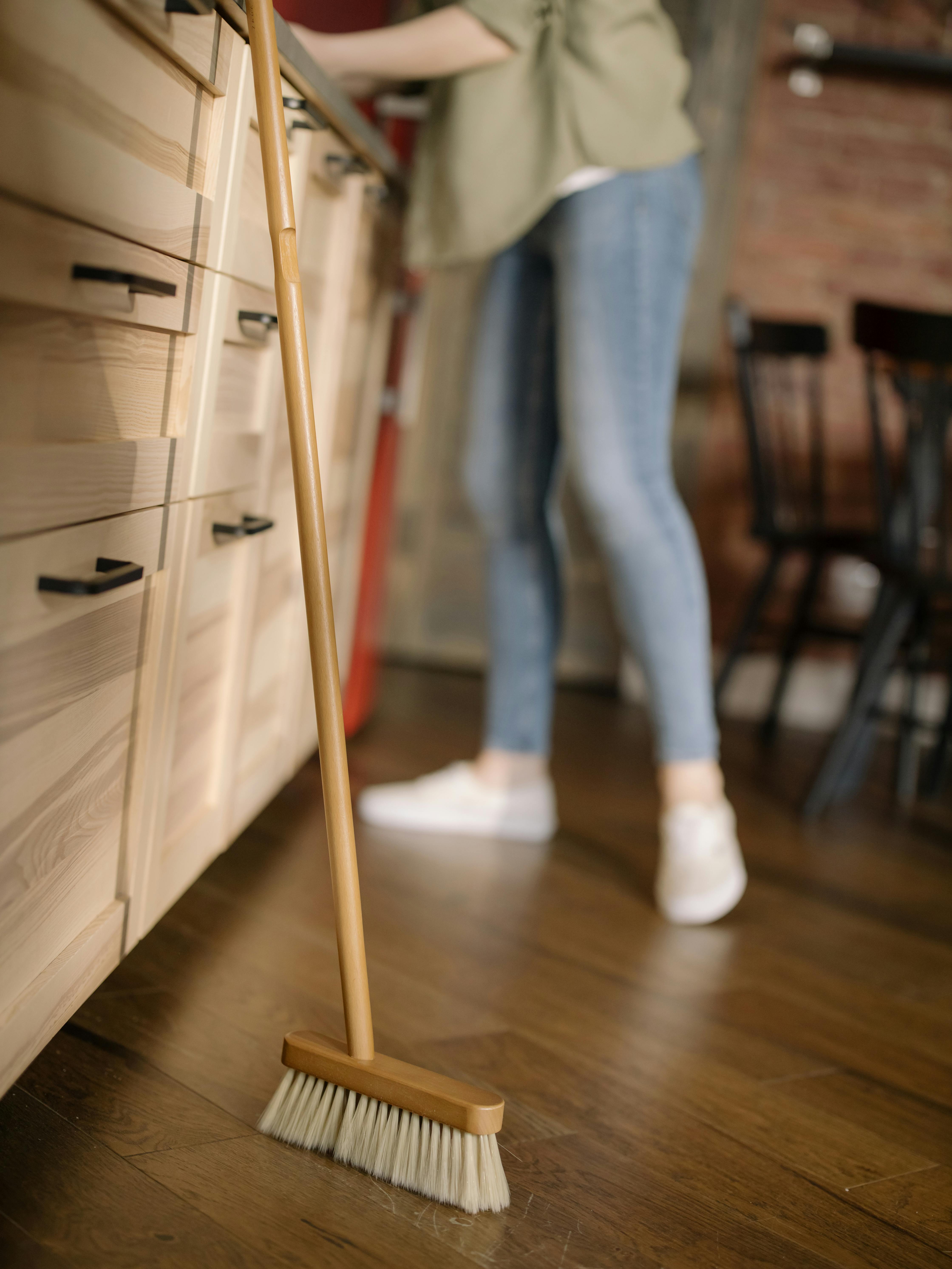 Person sweeping crumbs from wooden kitchen floor with broom as part of daily kitchen cleaning routine